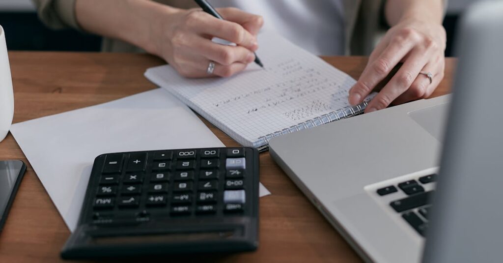 A woman writes financial calculations in a notebook, using a calculator and laptop at a wooden desk.